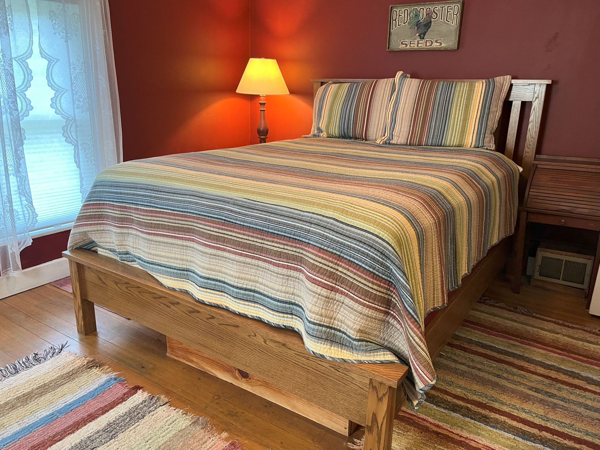 A bedroom with burgundy walls, a striped quilt on a wooden bed, and a window with white curtains.