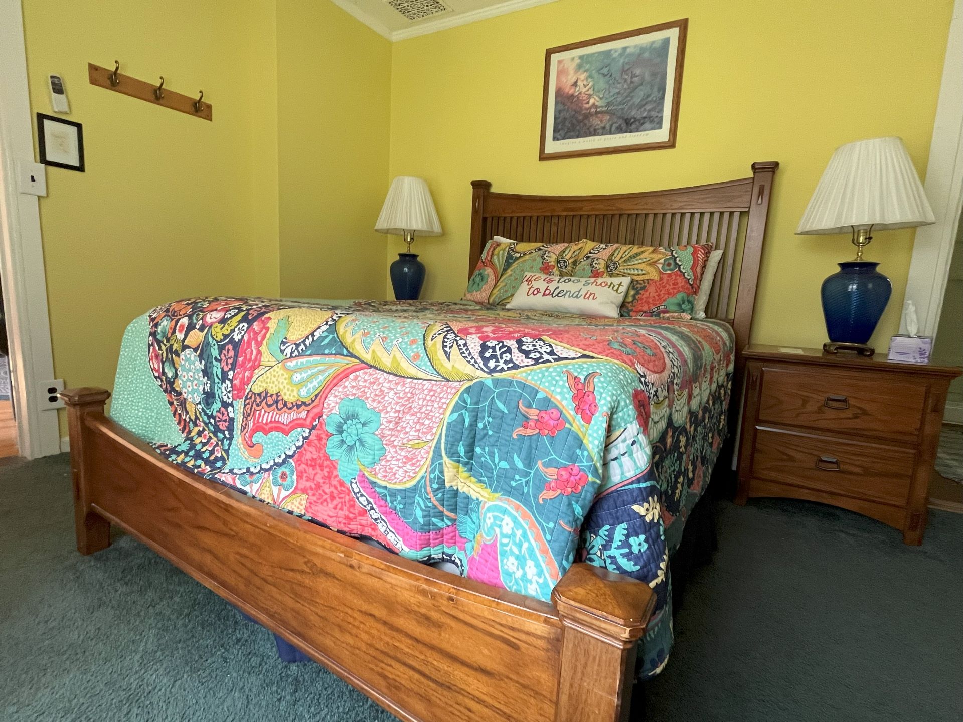 A bright bedroom with yellow walls, a colorful floral and paisley quilt, and a wooden bed frame.
