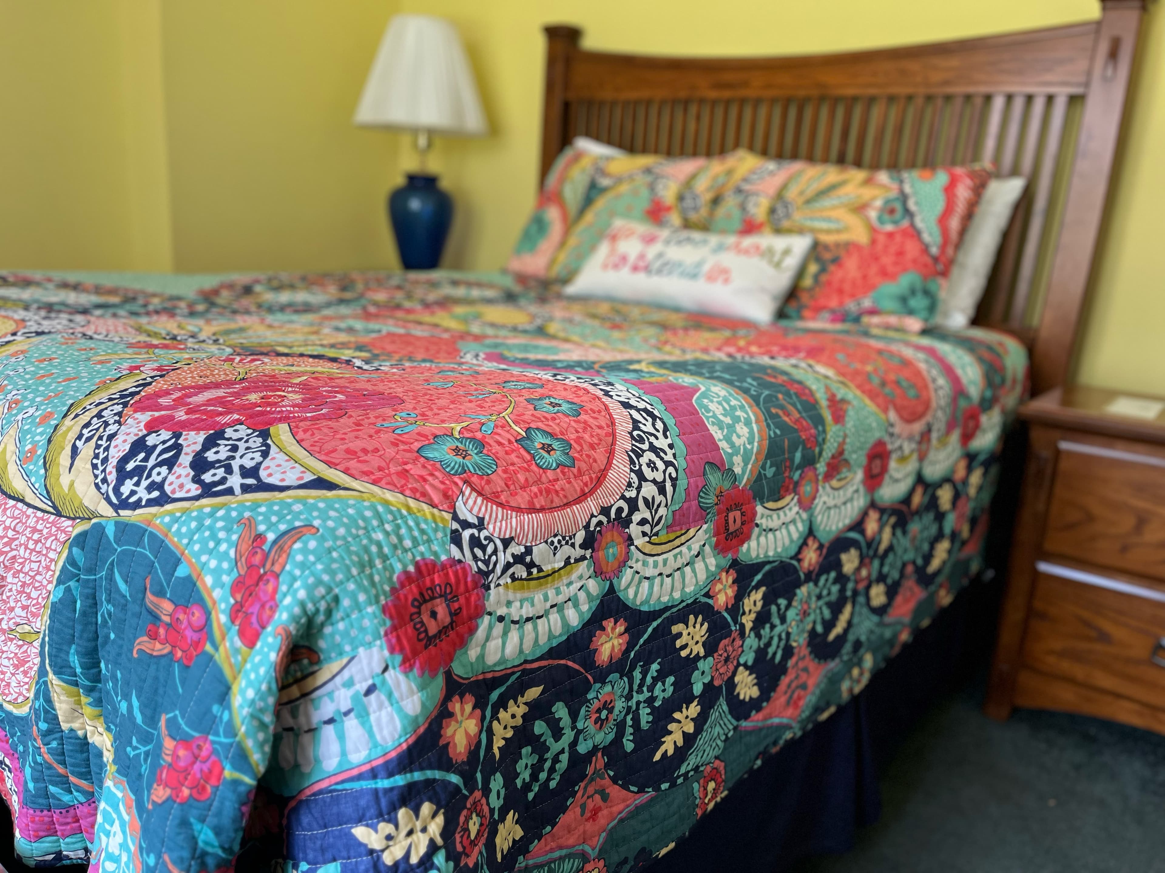 A bedroom featuring a bed with a colorful, floral-patterned quilt and a wooden headboard. There is a blue lamp on a nightstand next to the bed..