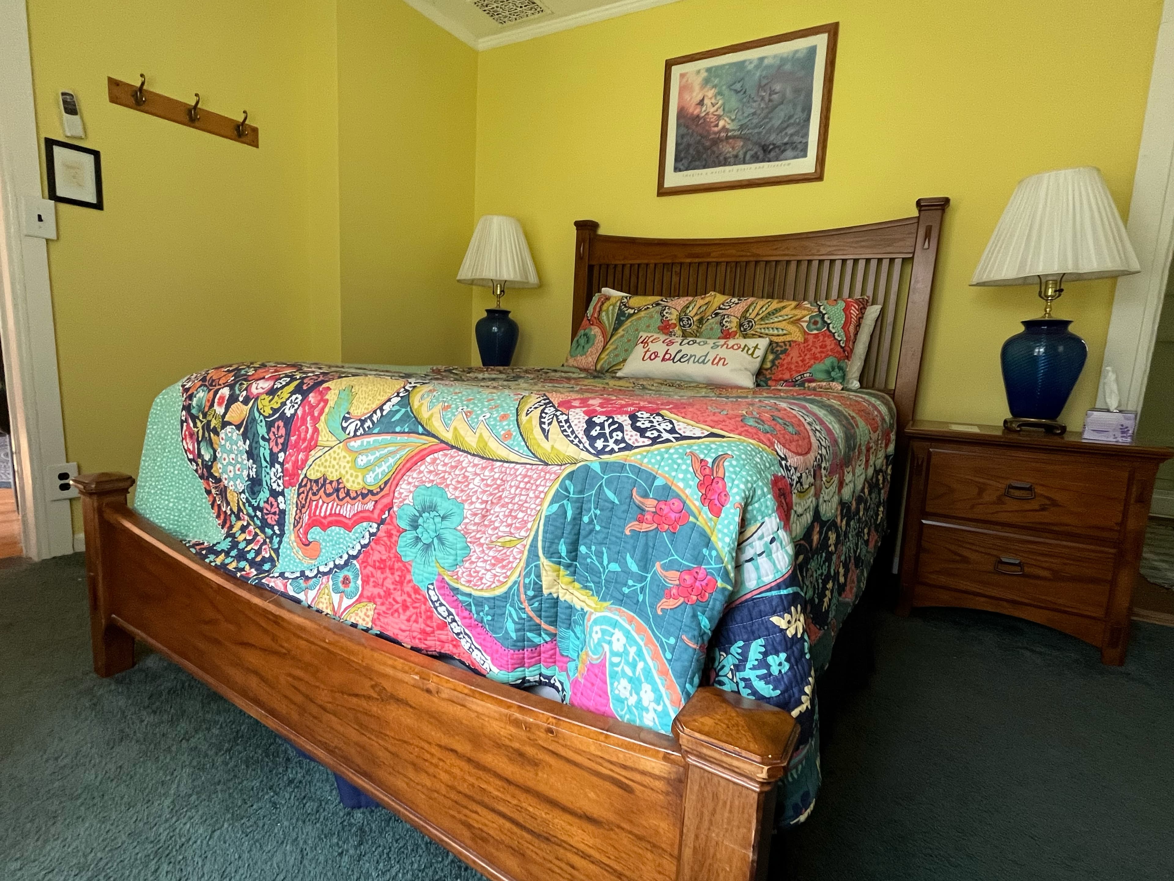 A colorful bedroom with yellow walls, featuring a wooden bed with a vibrant, paisley-patterned quilt. A matching lamp sits on each nightstand flanking the bed.