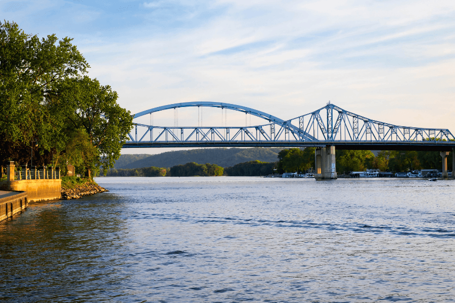 A blue bridge spans a calm river, flanked by greenery and a wooden bank.