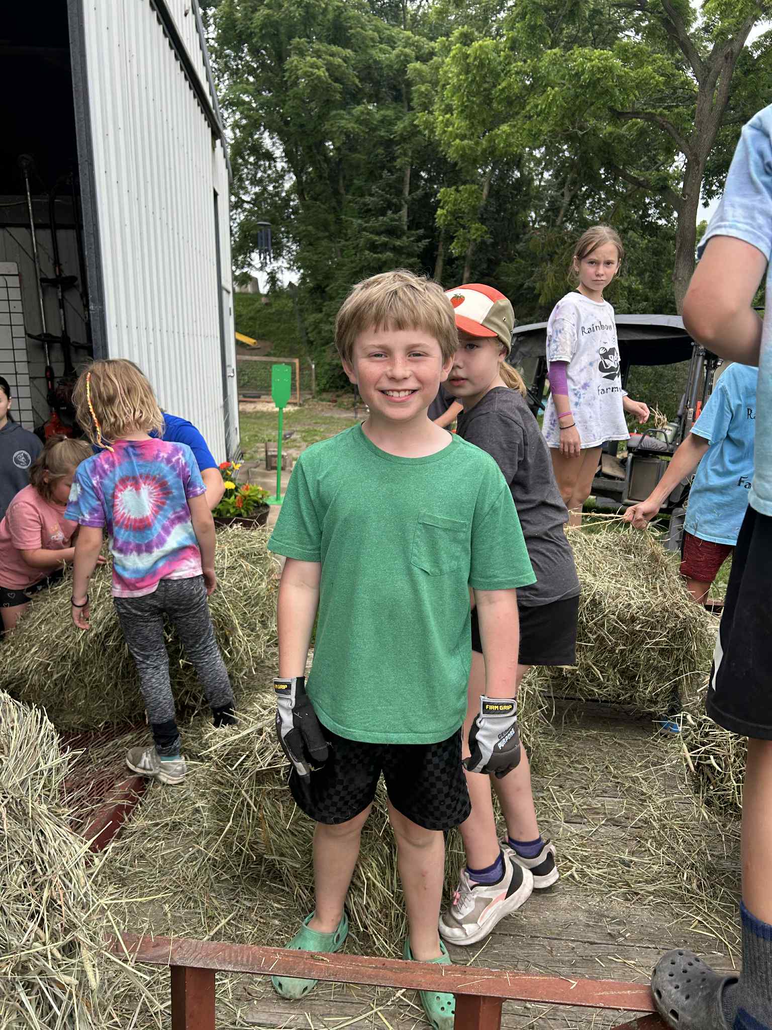A smiling boy in a green shirt and gloves stands among children on a farm, with hay bales around them.