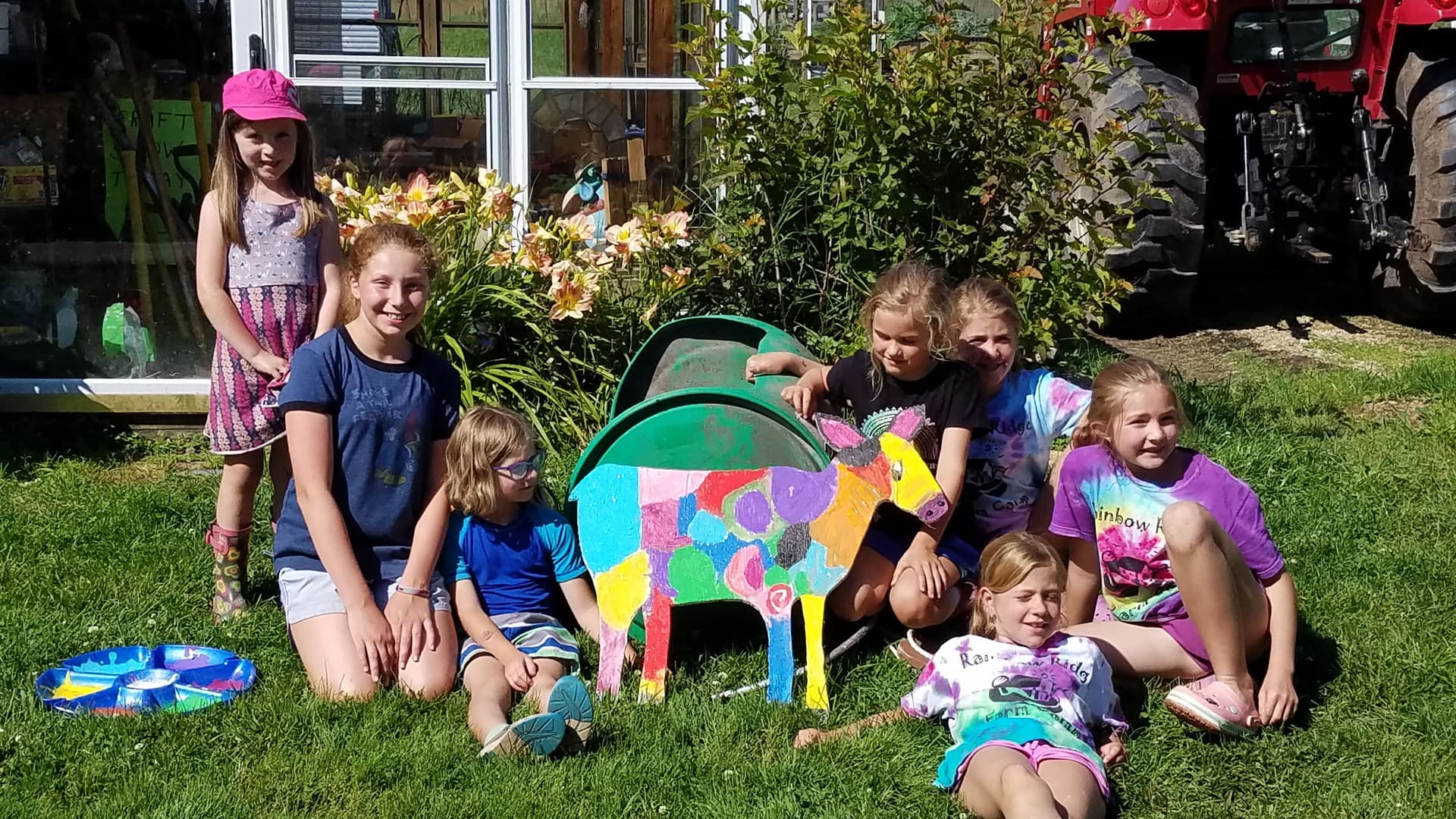 A group of six children poses with a colorful wooden cow statue in a garden.