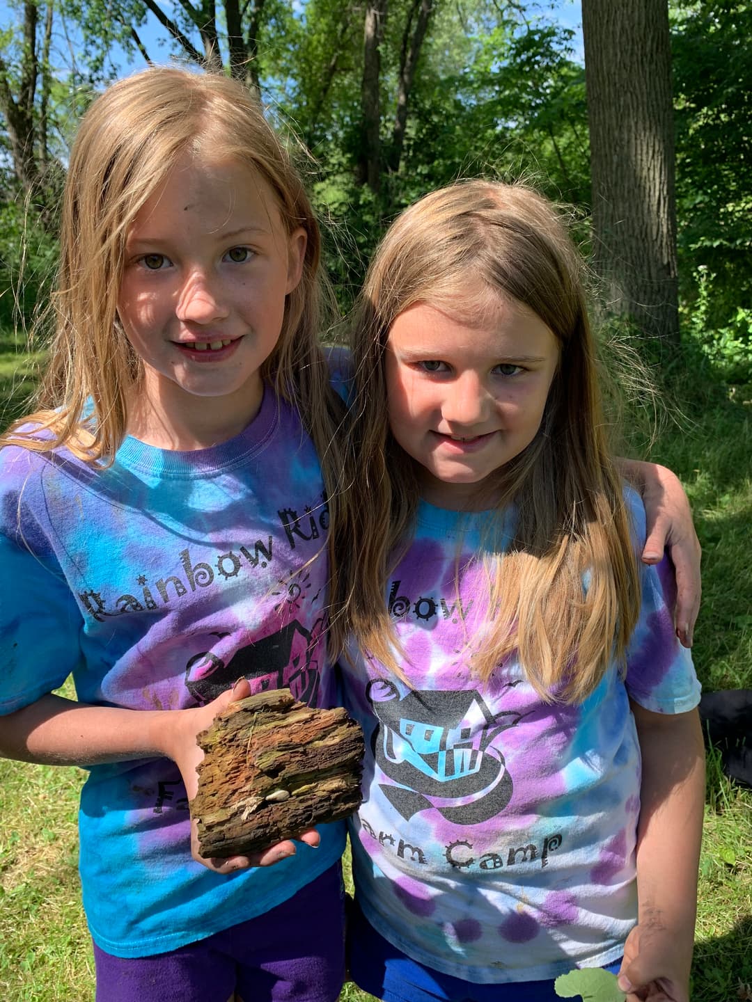 Two girls in tie-dye T-shirts hold a piece of wood while standing in a grassy area surrounded by trees.