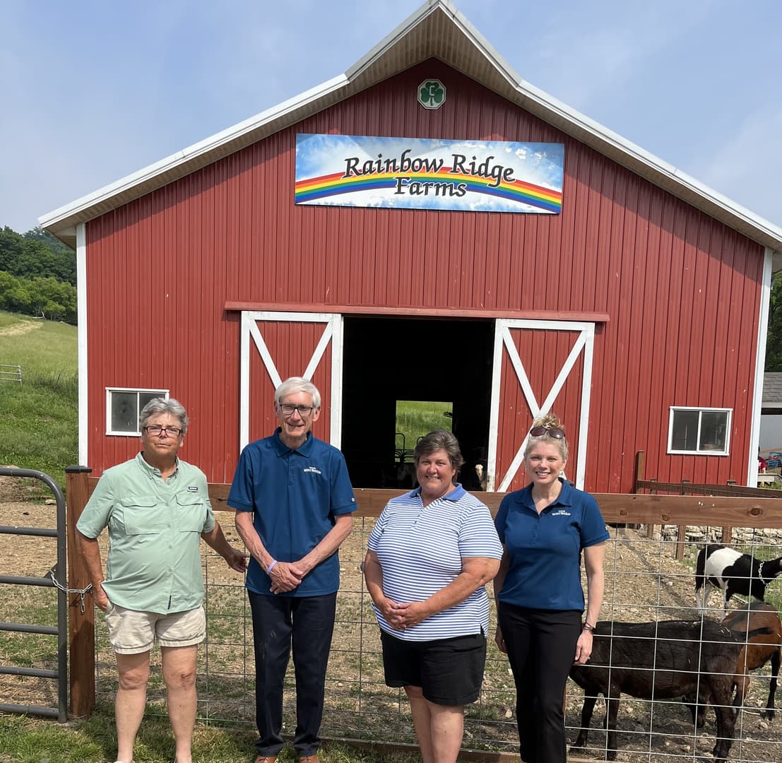 Four individuals stand in front of a red barn with a sign that reads "Rainbow Ridge Farms."