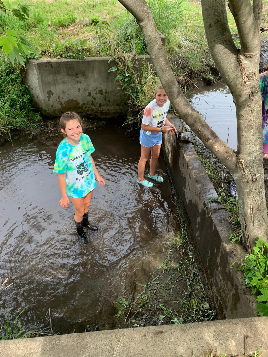 Two children in tie-dye shirts play in shallow water next to a concrete bank.